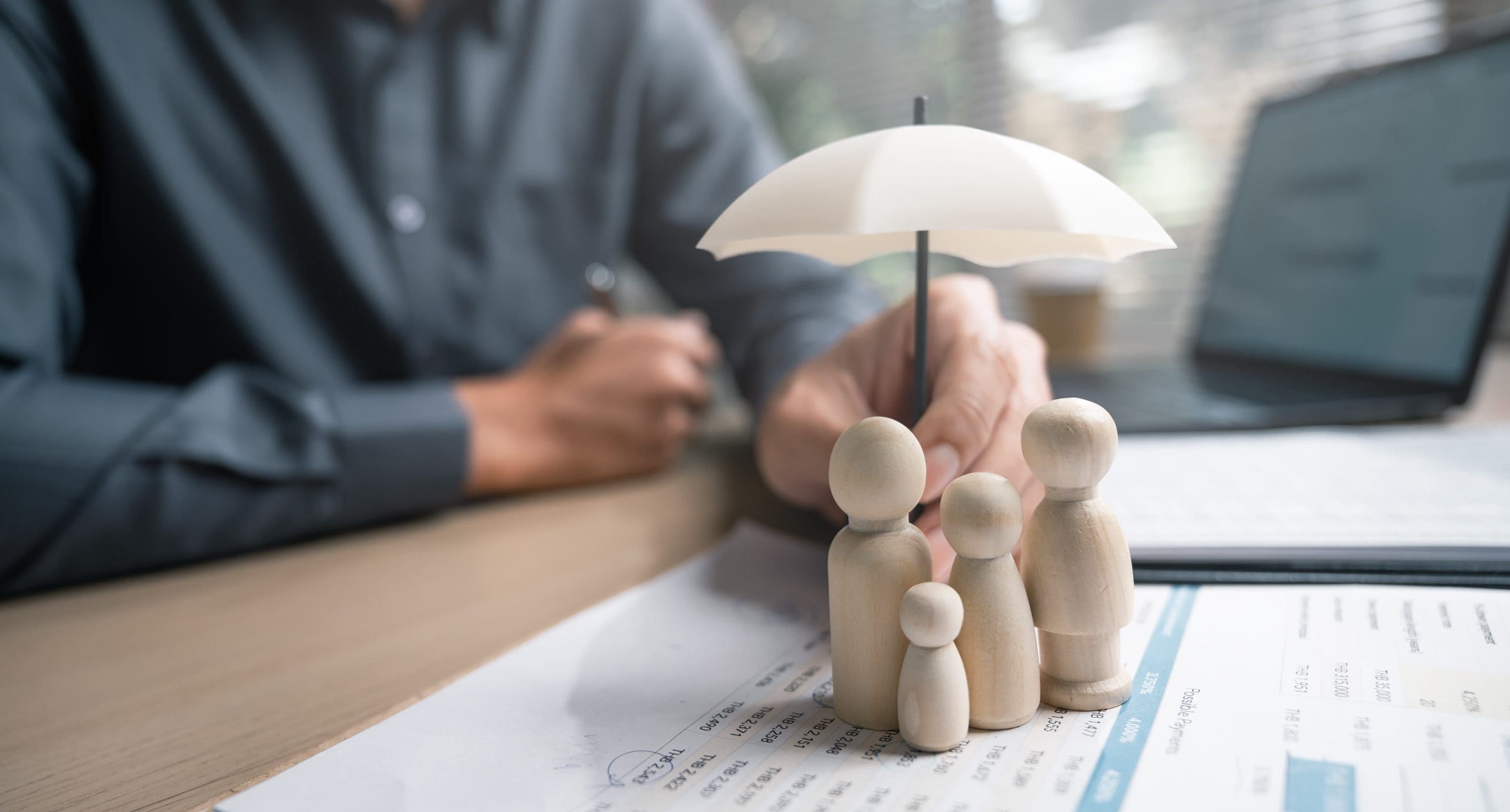 A person shields wooden family figures on documents with a small umbrella, illustrating liability insurance.
