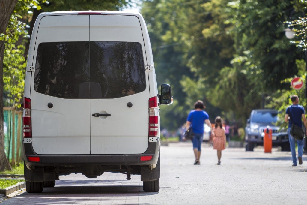 A white van is parked on a tree-lined street as people walk by on the sunny sidewalk in the background.
