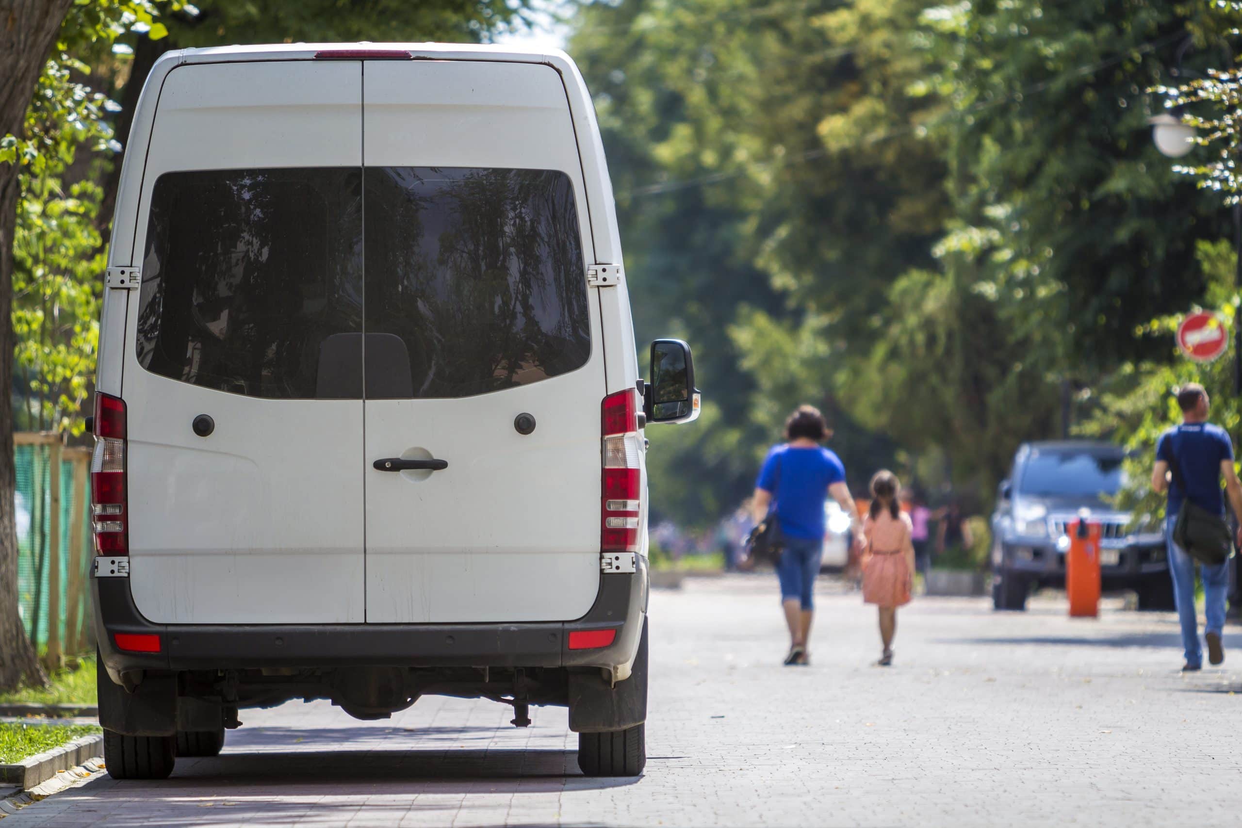 A white van is parked on a tree-lined street as people walk by on the sunny sidewalk in the background.