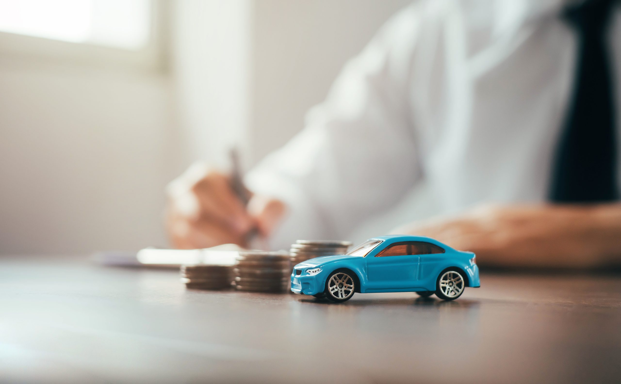 A blue toy car is next to stacked coins, with someone writing in the background, symbolizing auto finance.
