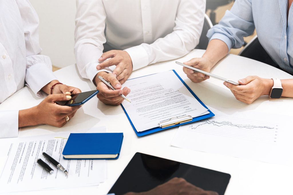 Three people at a table collaborate over documents, pens, a blue notebook, tablet, and smartphones.