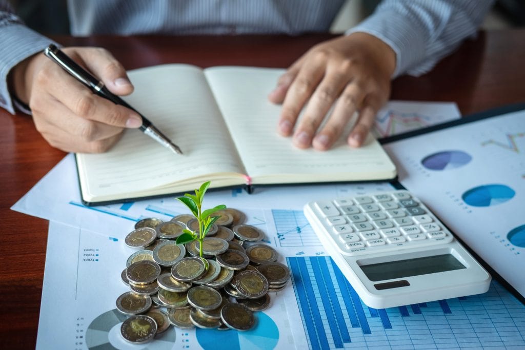 Person writing in a notebook with coins, calculator, and financial charts on a desk for budgeting.