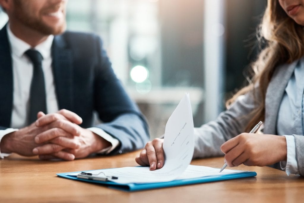 Two professionals in business attire sign a business document at a table during a meeting.