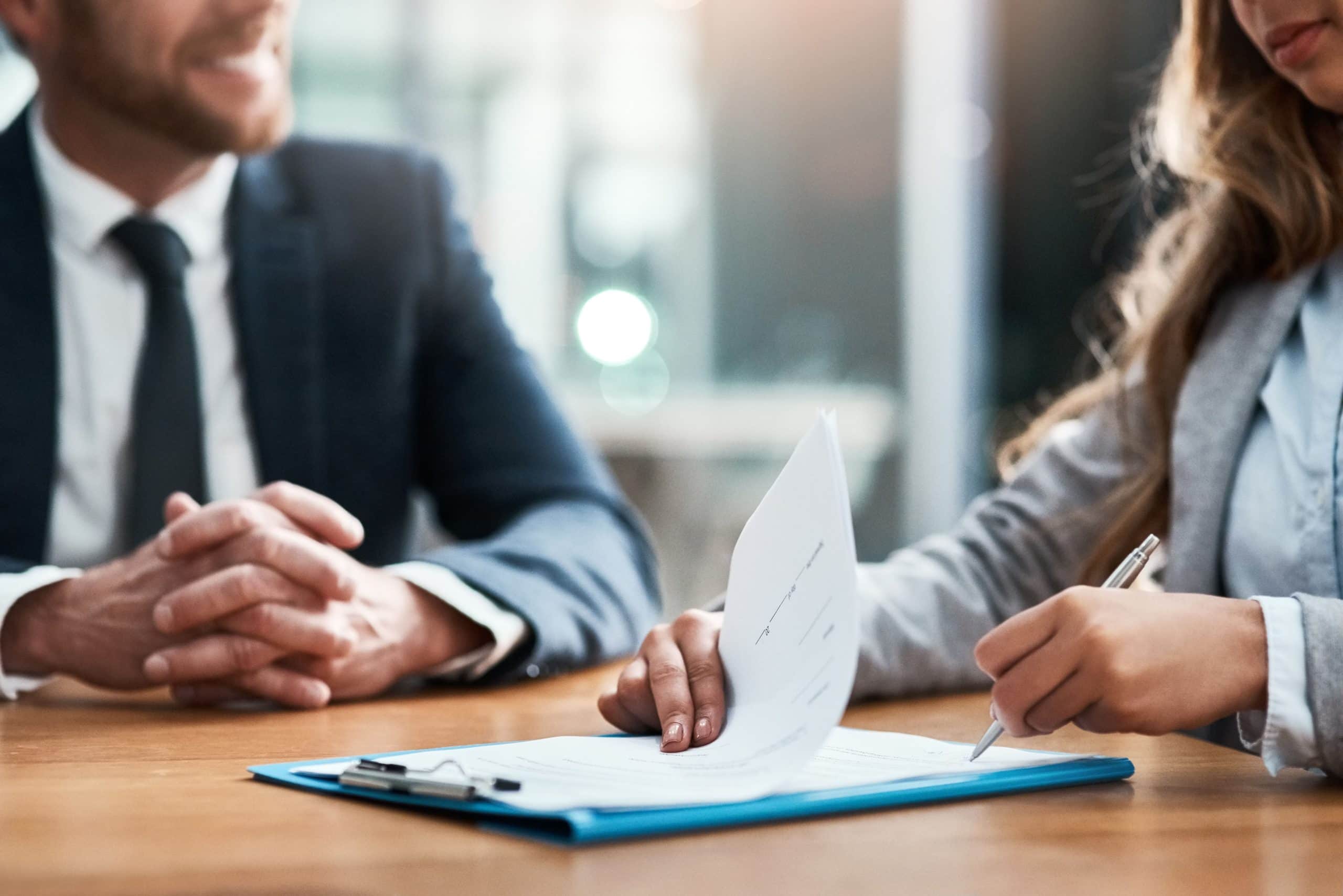 Two professionals in business attire sign a business document at a table during a meeting.