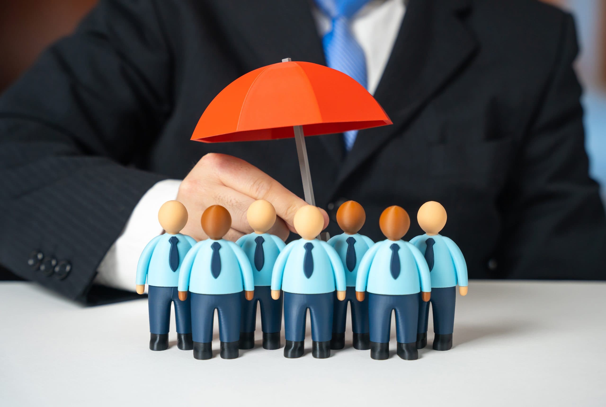 A man in a suit uses a red umbrella to protect small business figurines wearing ties from harm.
