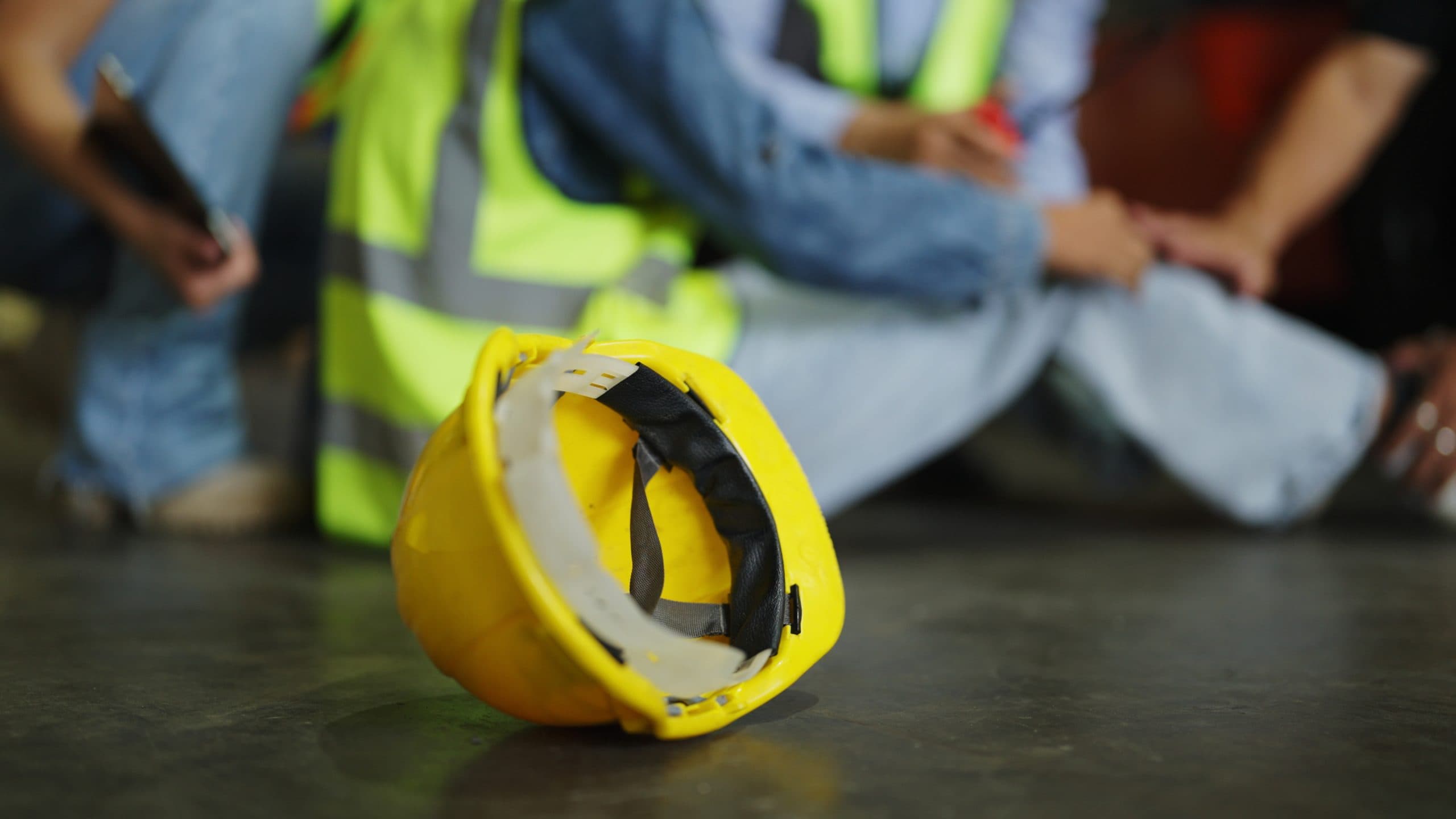 A yellow hard hat lies on the floor with a construction worker in the background holding an injured leg.