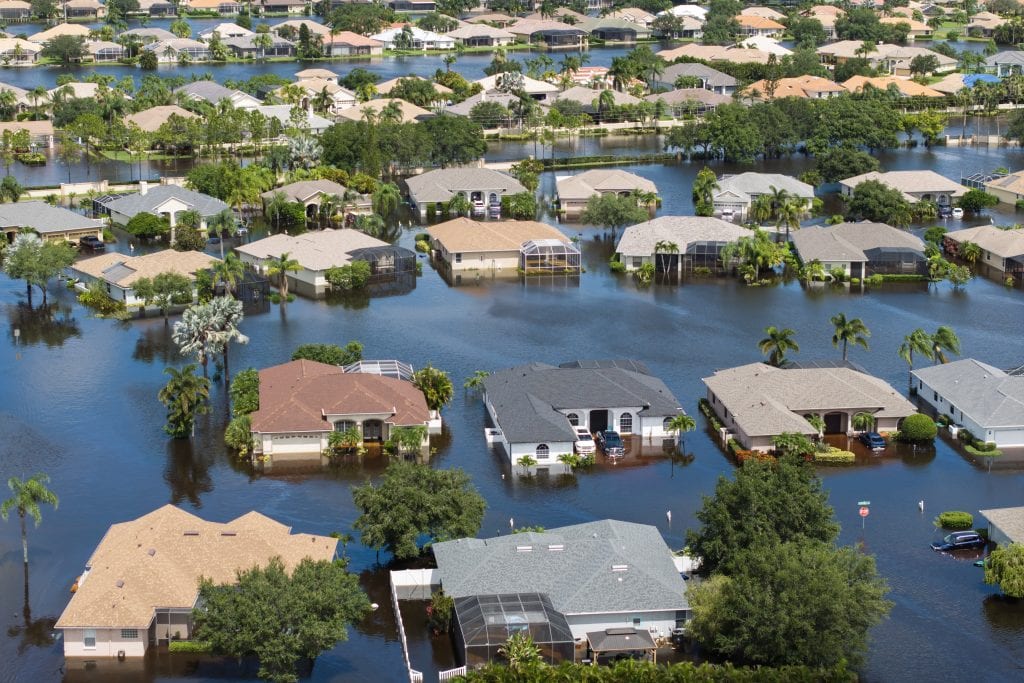 A suburban neighborhood is flooded, with water covering streets, houses, and trees after heavy rain.