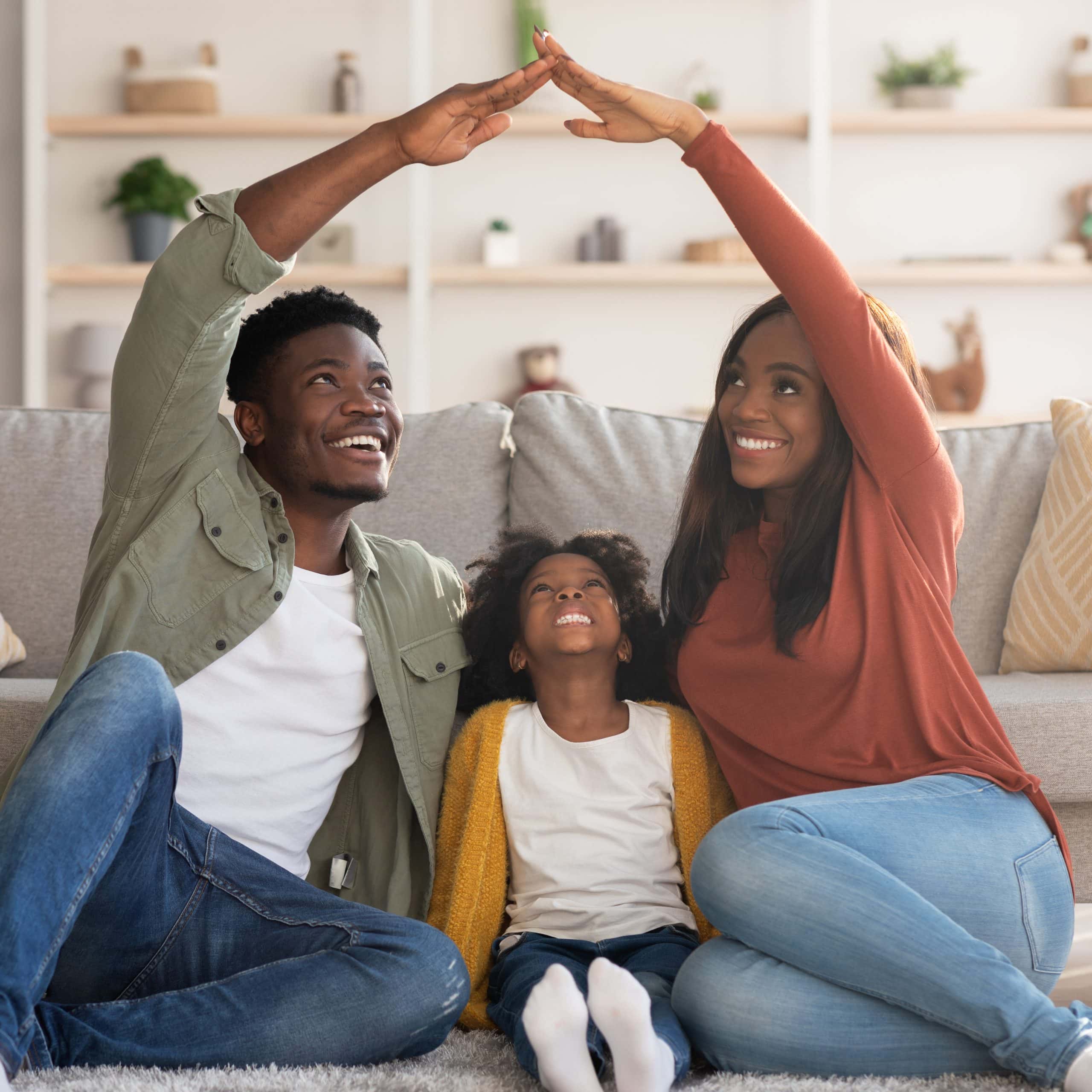 A smiling family with parents and child sits on a couch, parents forming a house shape with their hands.