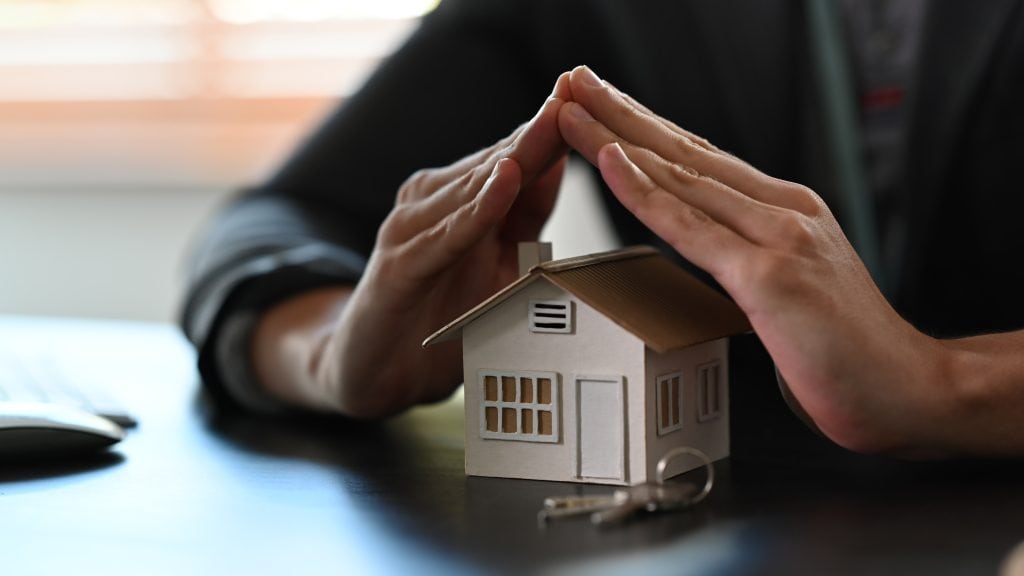 Hands form a roof shape over a small house model with keys, illustrating home protection and security.