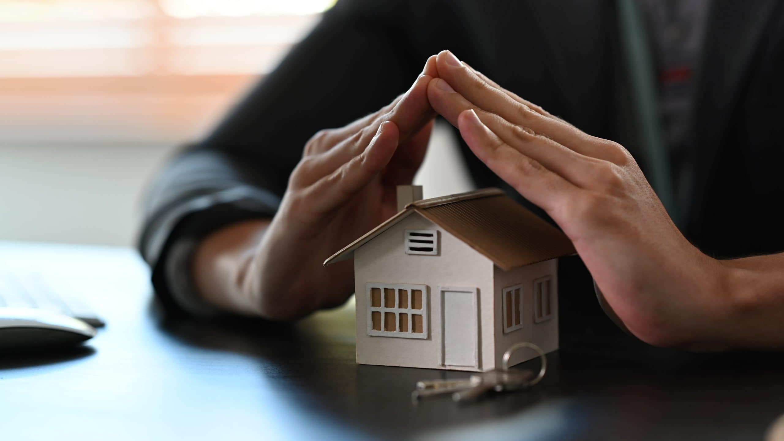 Hands form a roof shape over a small house model with keys, illustrating home protection and security.