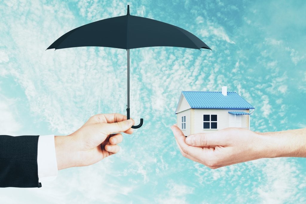 A hand holds an umbrella over a small model house, representing home protection and real estate insurance.