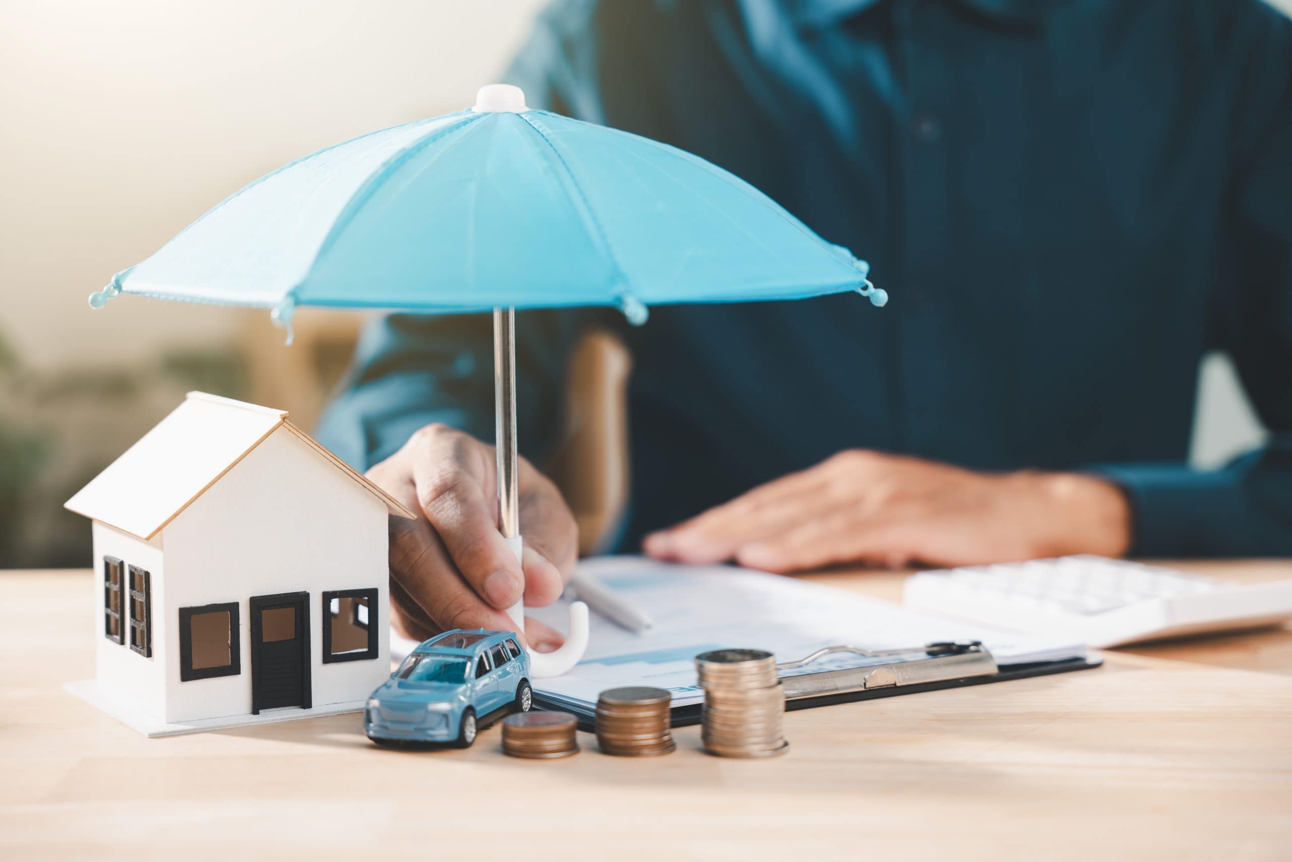 A person holds a blue umbrella over a house, car, and coins, representing insurance coverage and protection.