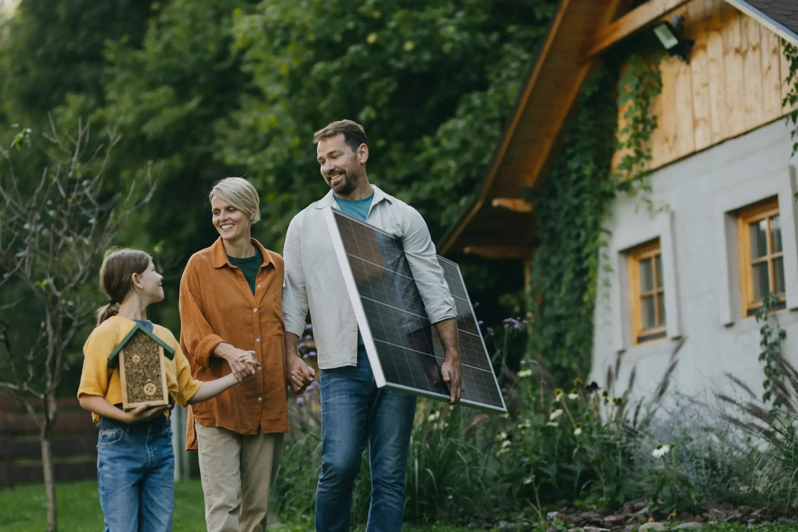 A family smiles outside their house, holding a birdhouse and solar panel in a lush green setting.