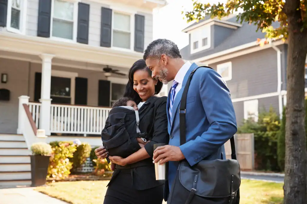 A smiling couple in business attire walks outside a house, carrying their baby in a front-facing carrier.