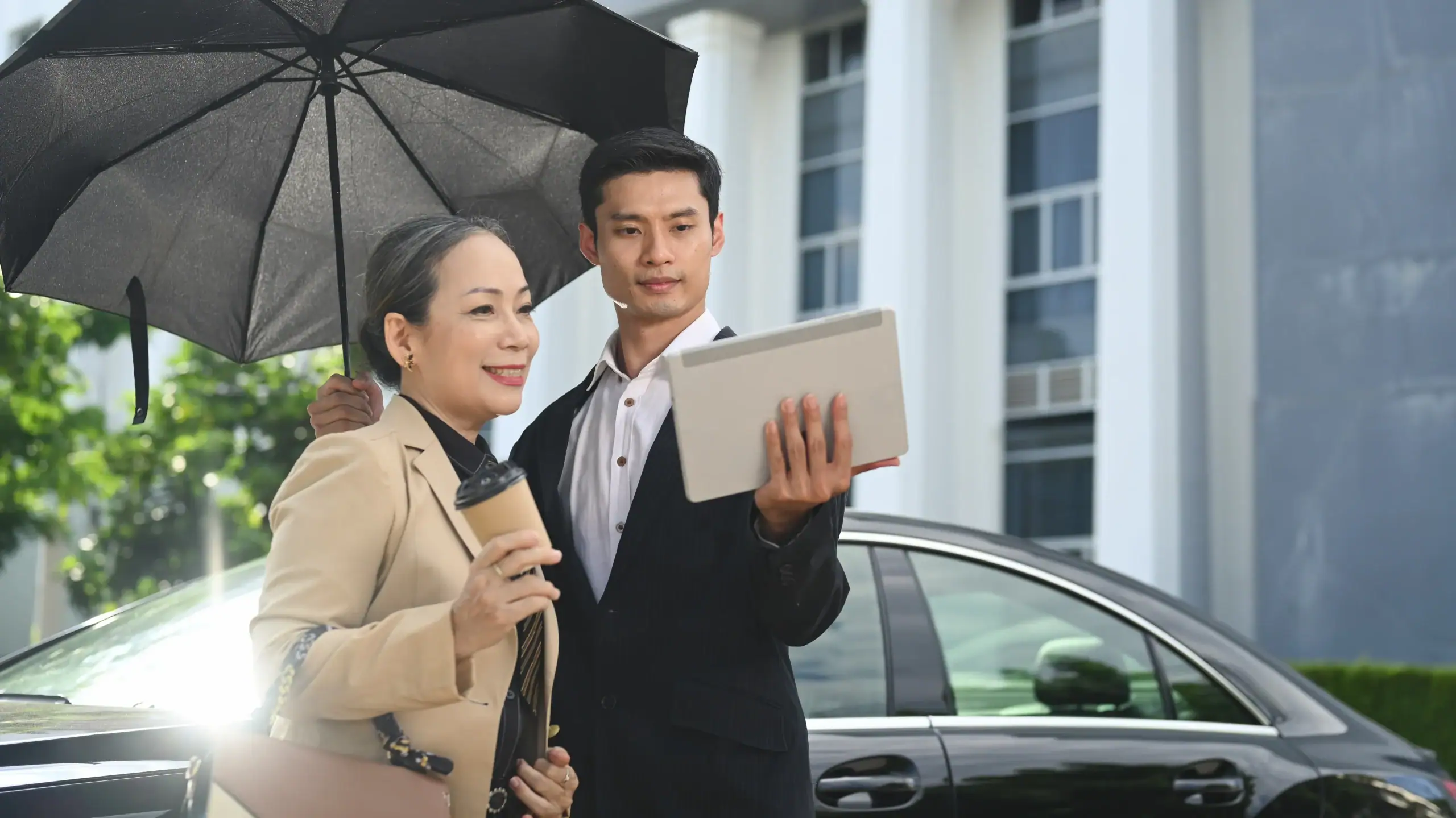 Two business professionals stand next to a car; one has an umbrella and tablet, the other holds coffee.