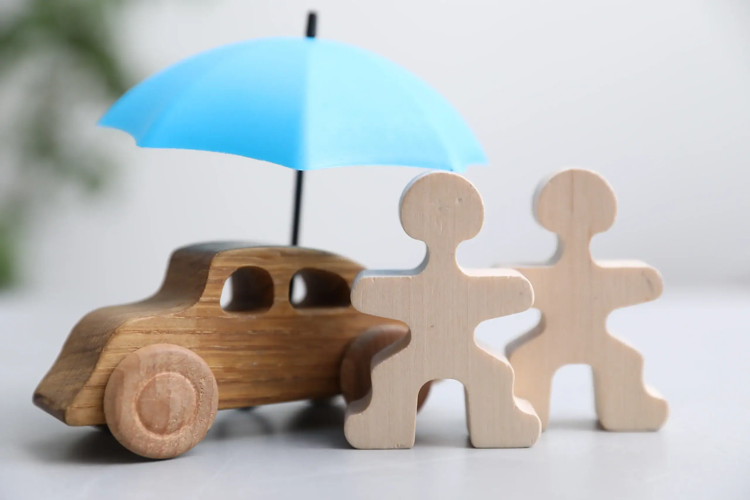 A toy wooden car and two figures sit under a small blue umbrella, symbolizing insurance or protection.